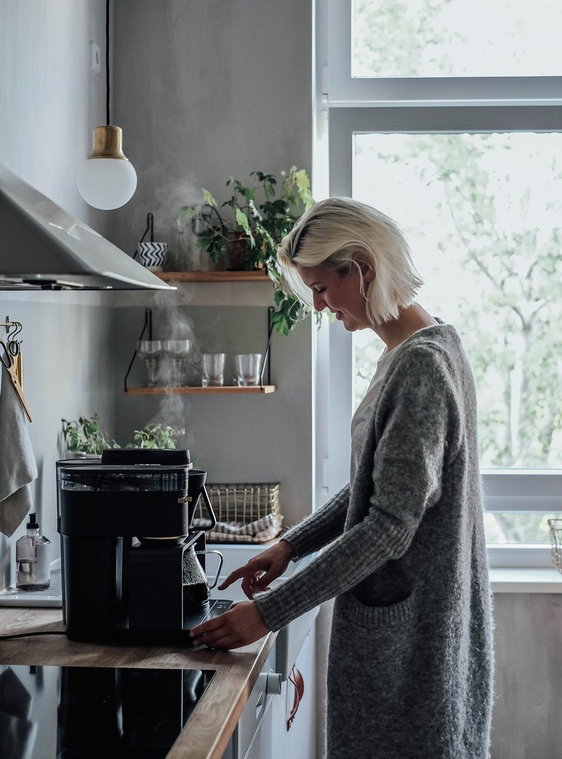 Blonde Frau steht an Küchenzeile mit elektrischer Pour Over Kaffeemaschine.
